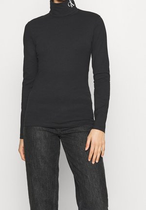 Personne portant un col roulé noir à manches longues avec le logo « ck » sur le col et un jean gris foncé, debout devant un fond clair uni.