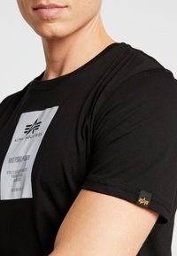 T-shirt en coton noir avec une étiquette rectangulaire blanche portant du texte et une petite étiquette jaune et noire sur la manche.