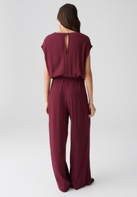 Bordeauxfarbener Jumpsuit mit kurzen Ärmeln, tailliertem Schnitt und einem Schlüsselloch-Detail am Rücken. Hergestellt aus leichtem Stoff; entspannter, weiter Schnitt.