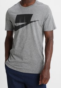 Šedé bavlněné tričko s černým grafickým motivem na přední straně, krátkými rukávy a kulatým výstřihem. Měkká textura a přiléhavý střih.