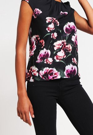 Haut noir sans manches à imprimé floral rose et blanc porté avec un pantalon noir, bras du mannequin levé près de la tête.