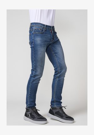 Mėlynos džinsinės siauro fasono kelnės su dėvėtu efektu, penkių kišenių dizainu ir kontrastingais siūlais, derinamos su pilkais aukštakulniais sportbačiais.