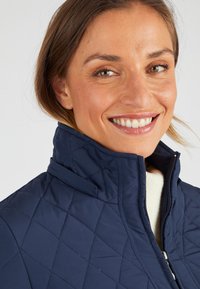 Marineblauwe gewatteerde jas met hoge kraag en rits, gemaakt van gladde stof. Voorzien van ruitvormige stiksels en een comfortabele voering.