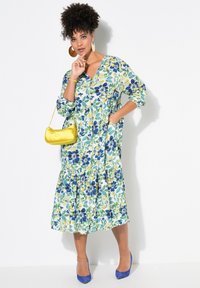 Abito con motivo floreale in tonalità blu, giallo e verde; lunghezza fino al ginocchio, scollatura a V, vestibilità ampia, vita arricciata, maniche lunghe. Borsa gialla, tacchi blu.