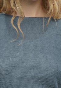 Pull en tricot bleu avec un col rond, présentant un motif texturé et un tissu léger et doux. Vue en gros plan du vêtement supérieur.