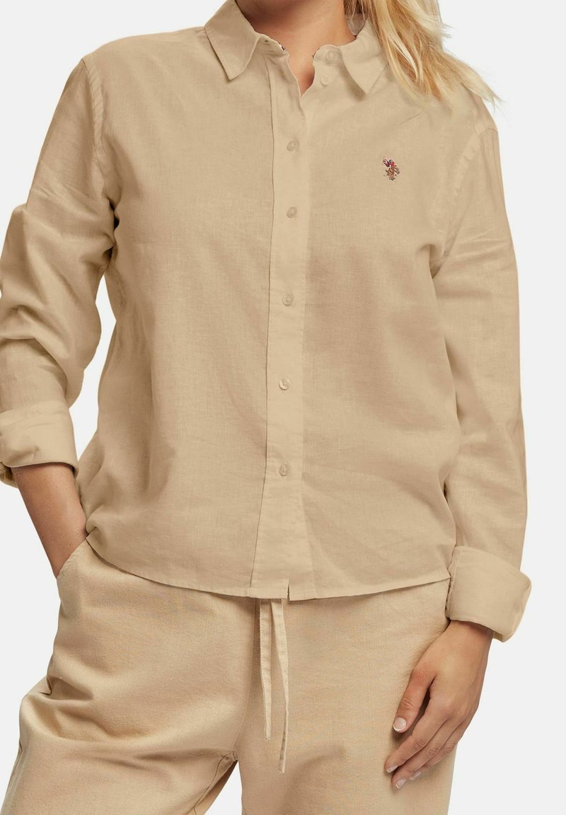 Camicia in lino beige chiaro con maniche lunghe, apertura con bottoni e un colletto leggero. Presenta un piccolo design ricamato sul petto.