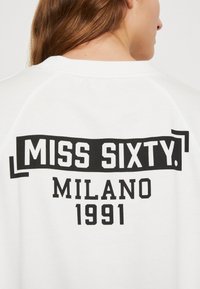 Valkoinen huppari, jossa on musta printti, jossa lukee "MISS SIXTY. MILANO 1991." Huomionarvoisia yksityiskohtia ovat pyöreä kaulus ja raglan-hihat.