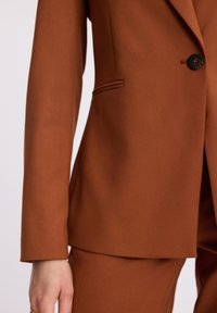 Brązowy slim fit żakiet z pojedynczym czarnym guzikiem i pasującymi spodniami, przedstawiający zbliżenie lewej ręki z dłonią noszącą złoty pierścionek.