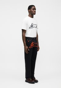 Λευκό βαμβακερό μπλουζάκι με σχέδιο μαύρης γάτας, συνδυασμένο με μαύρο τζιν που έχει σχέδιο κόκκινης γάτας. Το μοντέλο φορά καφέ παπούτσια.