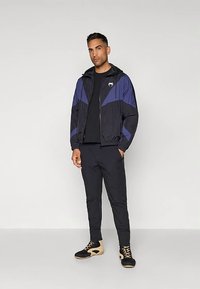 Mann står med hendene i lommene, iført svart og blå glidelåsjakke, svarte bukser, svart skjorte og svarte og beige høye sneakers.