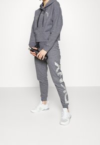 Šedá zkrácená mikina s kapucí a šňůrkami, doplněná o odpovídající tepláky s reflexním logem. Bílé sportovní boty dotvářejí outfit.