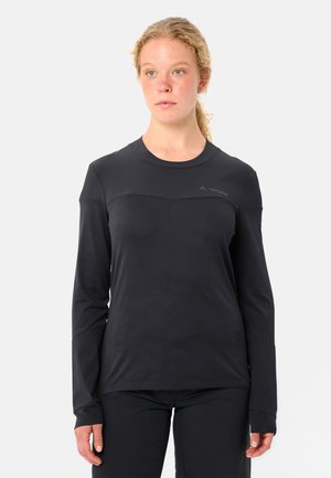 Junge Frau mit zurückgebundenem, leicht lockigem hellem Haar, trägt ein langärmliges schwarzes Sporttop und schwarze Hose, blickt mit neutralem Ausdruck nach vorne.