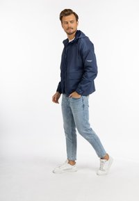 Giacca impermeabile blu navy con cappuccio, chiusura con zip e polsini elasticizzati. Indossata sopra una camicia bianca e abbinata a jeans azzurri chiari e sneaker bianche.