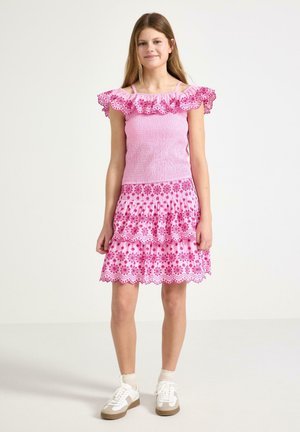 Pige stående iført en pink kjole med blomsterprint, flæset off-shoulder top og hvide sneakers mod en ensfarvet baggrund.