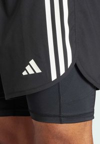 Bermudas deportivas negras con diseño de tres rayas blancas, que cuentan con una tela transpirable y un logotipo de Adidas en el dobladillo inferior.