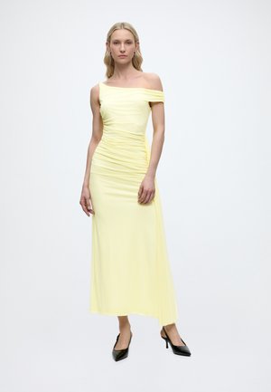 Žena plave kose nosi blijedo žutu midi haljinu na jedno rame sa naborima i crne cipele sa špicastim vrhom, stoji nasuprot bijele pozadine.