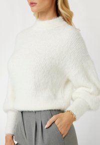 White, fluffy knit sweater with a high neck and loose sleeves, paired with gray pleated trousers. Bracelet accessory visible on wrist.