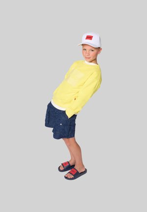 Boy leaning back, wearing a white cap, yellow sweatshirt, navy cargo shorts, and navy sliders with red logos on a gray background.