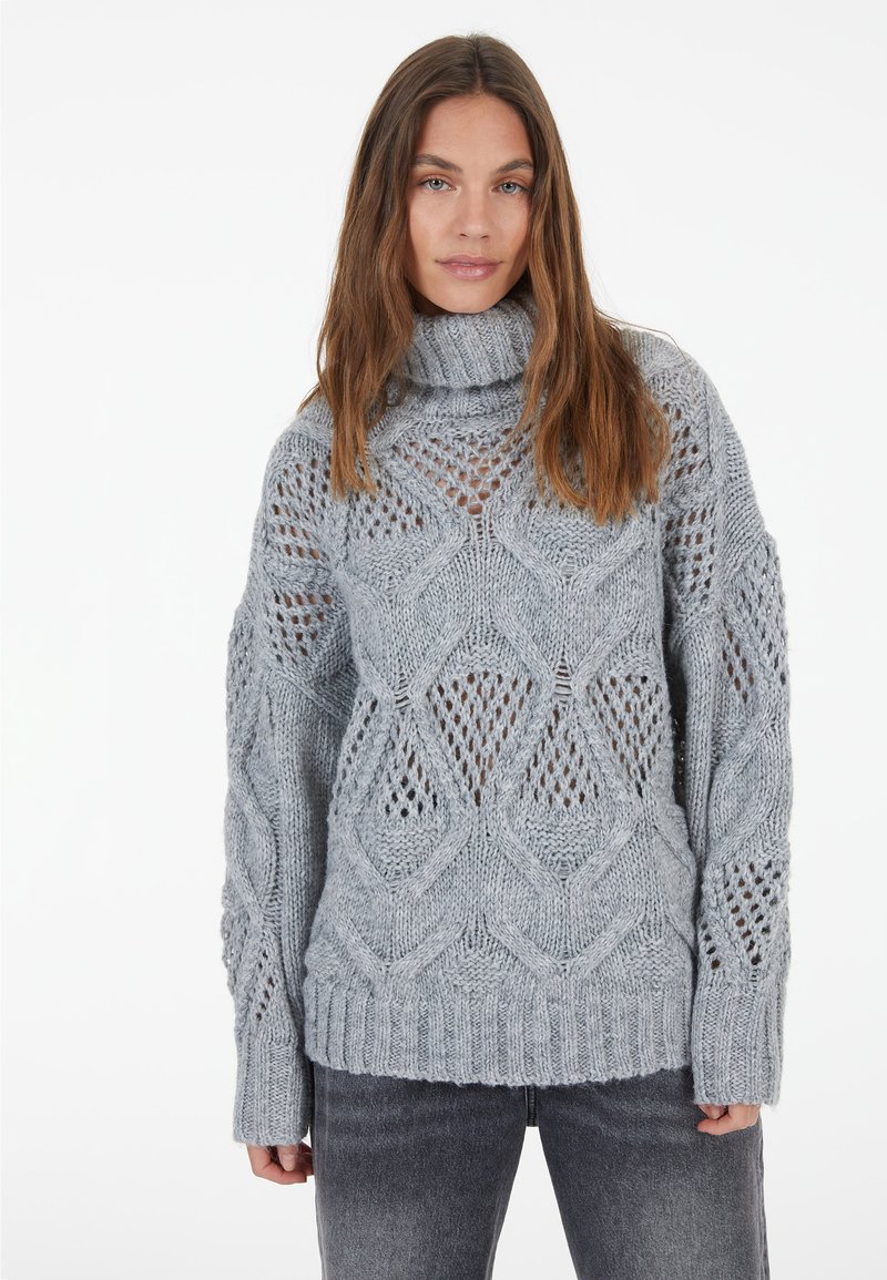 Hellgrauer Strickpullover mit hohem Kragen, der ein Lochmuster aufweist, lange Ärmel sowie gerippte Bündchen und Saum. Der strukturiertes Garn tritt hervor.