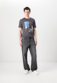 Grå bomull t-skjorte med grafiske trykk, parret med løstsittende mørke denimjeans og hvite joggesko, satt mot en enkel hvit bakgrunn.