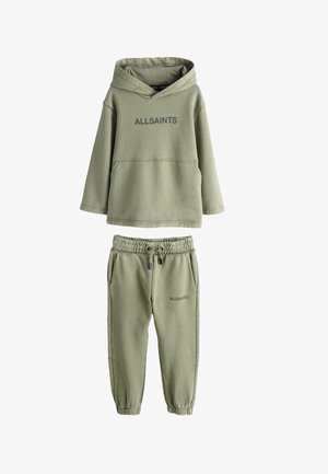 Jugendlicher in grünem ALLSAINTS Hoodie und Jogginghose, steht mit einer Hand in der Tasche und weißen Sneakers vor grauem Hintergrund.