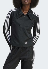 Casaco atlético preto com riscas brancas nas mangas, apresentando um colarinho e bolsos com fecho éclair. Logótipo do fabricante visível no peito.