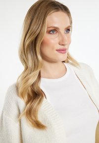 Witte gebreide cardigan over een witte top, met een ronde halslijn. Het haar is golvend en lichtblond, met blauwe ogen en neutrale make-up.