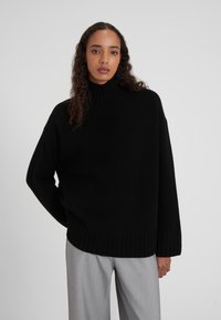 Pull en maille noir oversize avec col roulé et bords côtelés, offrant une coupe décontractée et un tissu texturé. Associé à un pantalon gris clair.