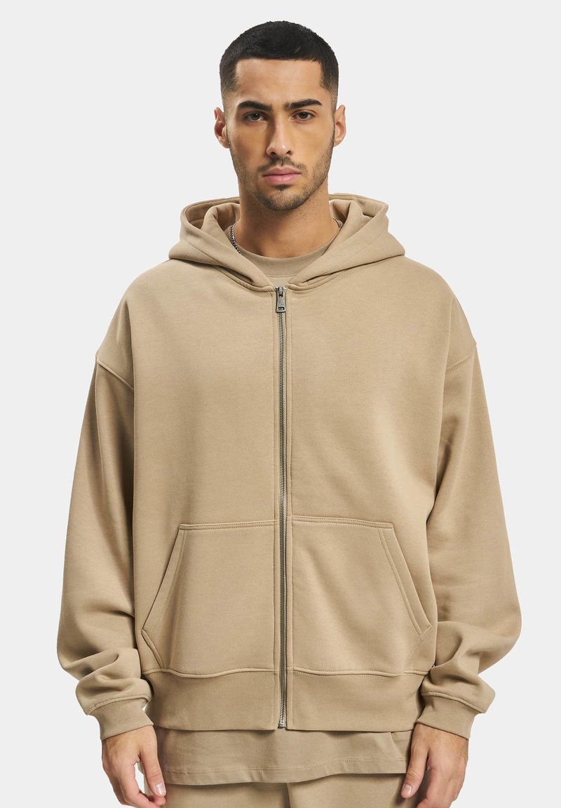 Haut zippé beige en matériau doux, avec une capuche spacieuse, des poches avant et une coupe décontractée. Design simple avec des poignets côtelés.