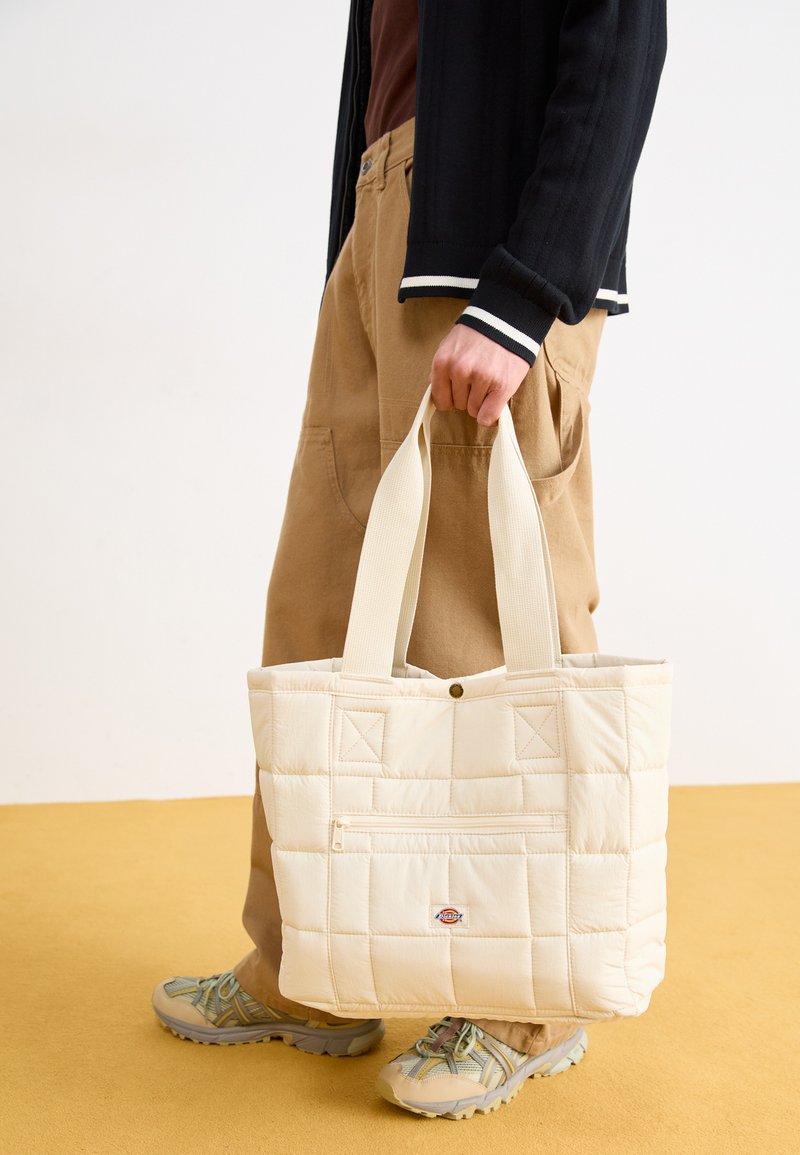 Gepatchte cremefarbene Tragetasche mit zwei Griffen, einer Vordertasche und einem Logopatch; kombiniert mit beigen Cargo-Hosen und neutralen Sneakers.
