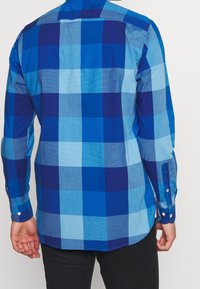 Hombre con una camisa de cuadros azul de manga larga de espaldas a la cámara, combinado con pantalones negros.