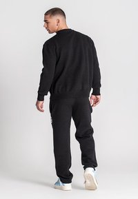 Junger Mann, der von hinten geht, trägt einen schwarzen Sweatshirt und Sweathosen sowie weiße und blaue Turnschuhe vor einem grauen Hintergrund.