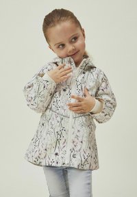 Giacca bianca con un motivo floreale in viola e verde. Presenta una chiusura con zip, cappuccio e polsini elasticizzati. Indossata con jeans blu chiaro.