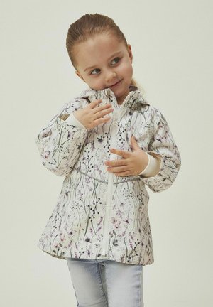 Giacca bianca con un motivo floreale in viola e verde. Presenta una chiusura con zip, cappuccio e polsini elasticizzati. Indossata con jeans blu chiaro.