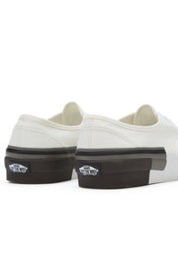 White canvas shoes with a low-profile design, featuring black rubber soles, logo accents, and stitched detailing on the upper.