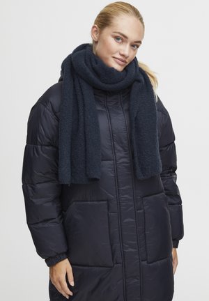 Tumesinise puffer-kate, millel on tikitud tekstuur, esine sakiga ja küljetaskudega, koos tumeda sinise villase salliga, mis on kaela ümber korda keeratud.