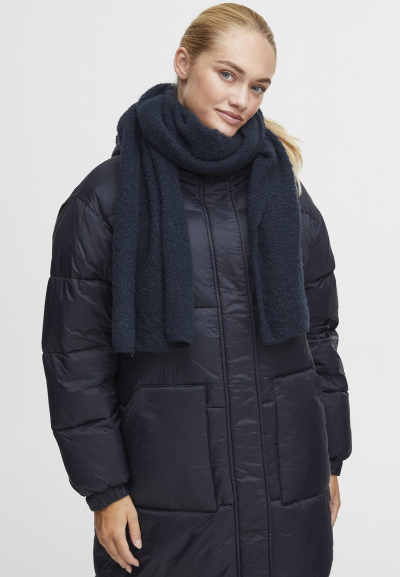 Navy puffer coat with quilted texture, featuring a front zipper and side pockets, paired with a dark blue woolen scarf wrapped around the neck.