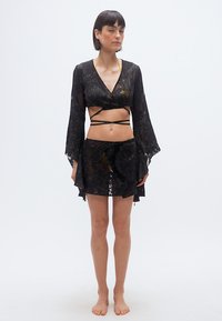 Γυναίκα που στέκεται ξυπόλητη φορώντας μαύρο δαντελένιο crop τοπ με μακριά μανίκια και ταιριαστή μαύρη δαντελένια μίνι φούστα σε απλό λευκό φόντο.