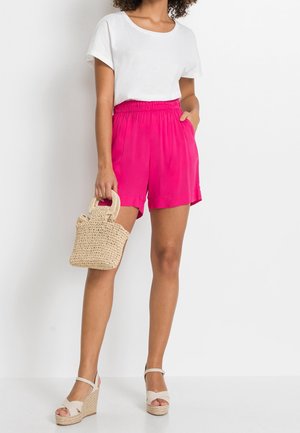 Des shorts légers et roses avec une taille élastique, associés à un haut blanc à manches courtes. Porte un petit sac tissé. Sandales compensées beiges.