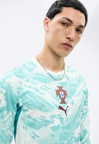 Νεαρός άνδρας με κοντά σκούρα μαλλιά και φακίδες, φοράει φανέλα ποδοσφαίρου της Πορτογαλίας σε ανοιχτό γαλάζιο και λευκό με μοτίβο, με το λογότυπο της Puma και ασημένια αλυσίδα.