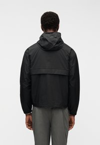 Άνδρας με μαύρο μπουφάν με κουκούλα και γκρίζο παντελόνι, εμφανιζόμενος από πίσω μπροστά σε απλό λευκό φόντο.