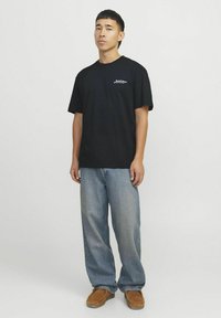 Camiseta de algodón negra de mangas cortas con un pequeño logo, combinada con jeans sueltos de color azul claro y zapatos marrones sin cordones.