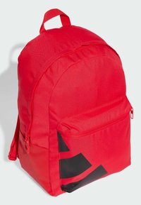 Mochila roja de tela con un bolsillo lateral con cremallera, que presenta un acento geométrico negro en la parte frontal inferior. Forma redondeada y asa superior.