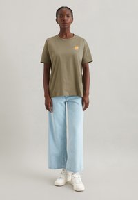 Olivgrünes Baumwoll-T-Shirt mit einem Sonnenmotiv, kombiniert mit hellblauen weit geschnittenen Jeans und weißen Sneakern, vor einem neutralen Hintergrund stehend.