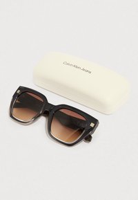 Óculos de sol Calvin Klein Jeans com armação grossa preta que se mistura num degradê transparente; lentes em degradê castanho; inclui estojo de cor creme.