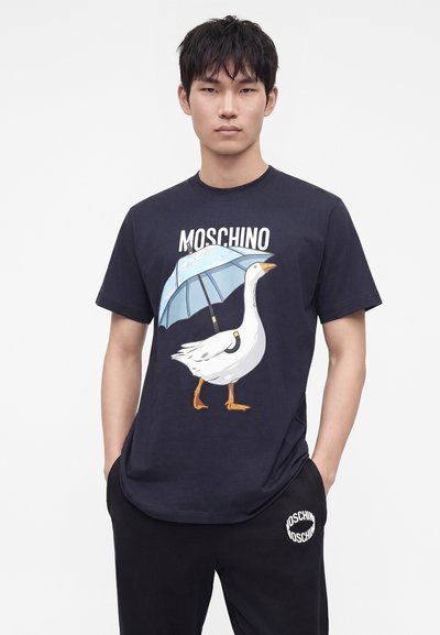 Νέος άνδρας φοράει μπλε μπλούζα Moschino με γραφική απεικόνιση μιας χήνας που κρατάει μπλε ομπρέλα και μαύρο παντελόνι με το λογότυπο Moschino.