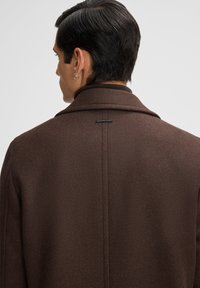 Bruine wollen jas met een gesneden kraag, voorzien van een subtiele visgraatstructuur en een merklabel op de bovenrug.