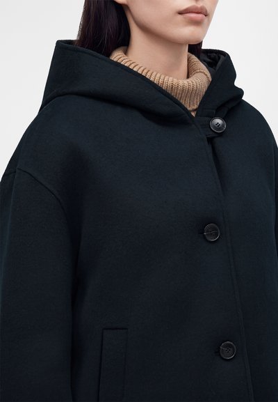 Personne portant un manteau noir à capuche avec trois grands boutons et un pull à col roulé côtelé beige en dessous.
