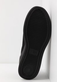 Semelle en caoutchouc noire d’une chaussure avec des rainures horizontales et un motif circulaire près du talon, présentée sur fond blanc.