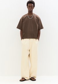 Braun oversized T-Shirt mit kurzen Ärmeln, lockere cremefarbene Hose und schwarze Flip-Flops. Eine Ketten-Halskette als Akzent. Einfaches, entspanntes Design.